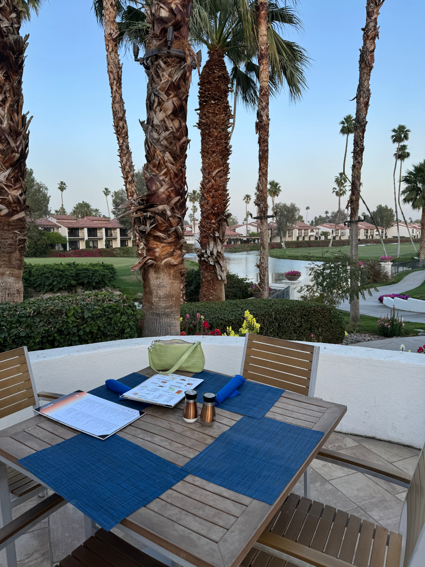 Outdoor dining table set with notebooks, overlooking a pond with palm trees in the background