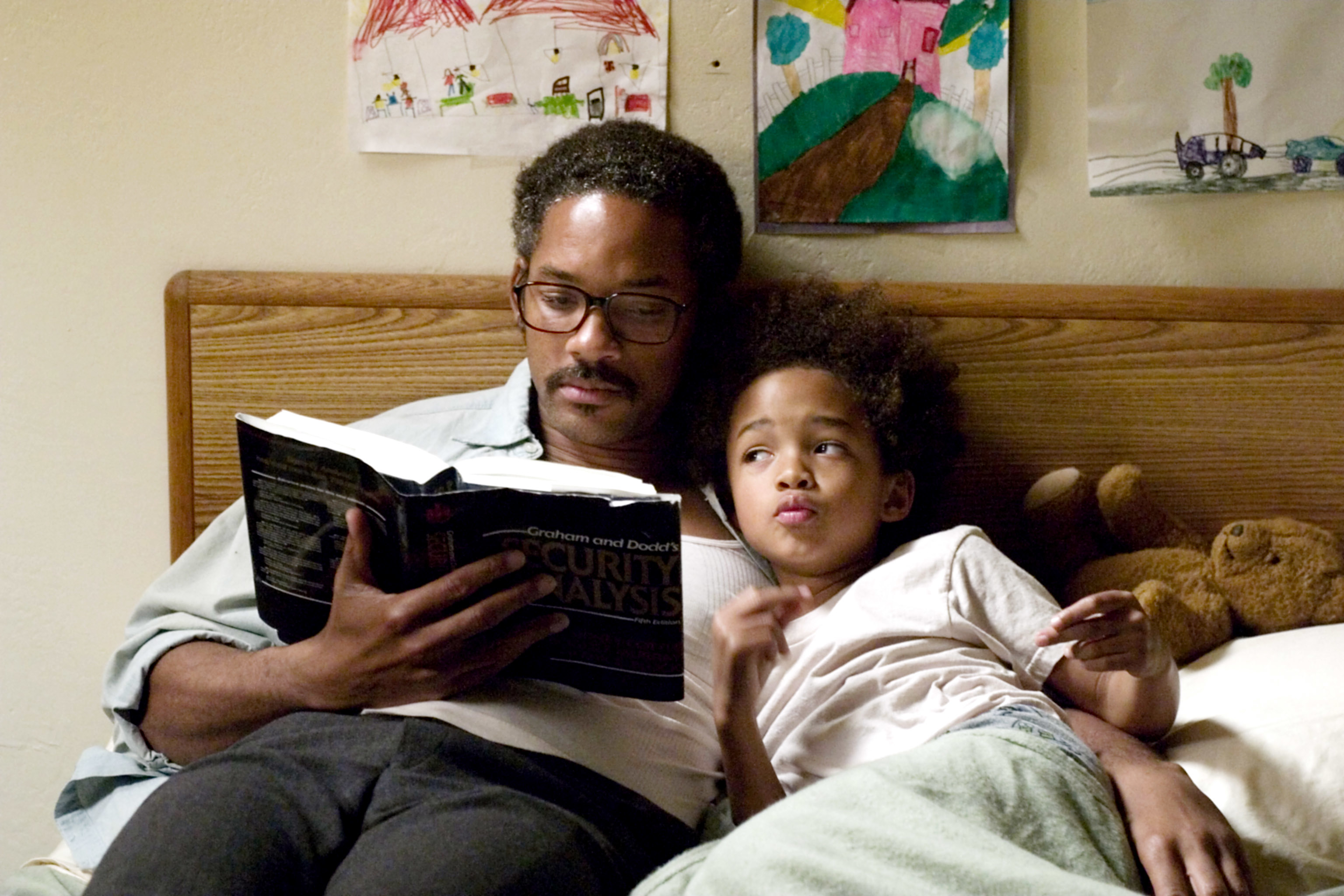 Man and child relax in bed, man reads book, child listens. They are TV characters in a domestic scene