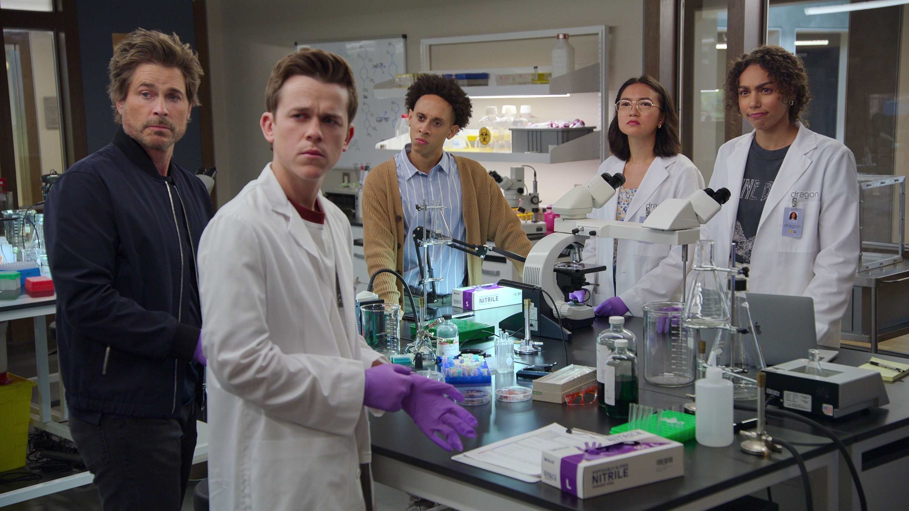 Five characters in a lab from a TV show, standing by equipment, looking seriously at the camera