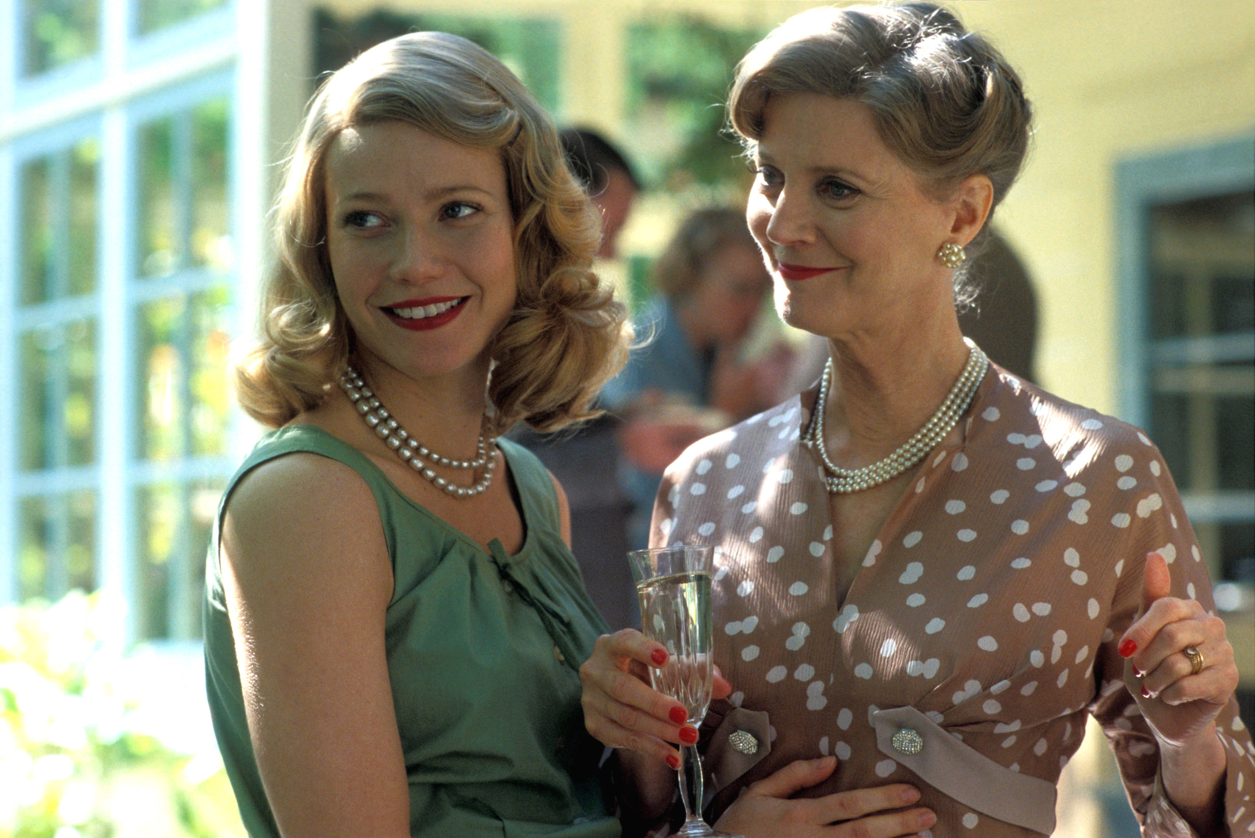 Actresses in period attire with pearls, one in a polka dot dress, holding champagne, in a TV or movie scene