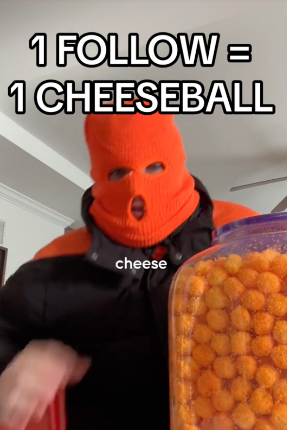 Person in orange ski mask, holding a sign, standing next to a container of cheeseballs