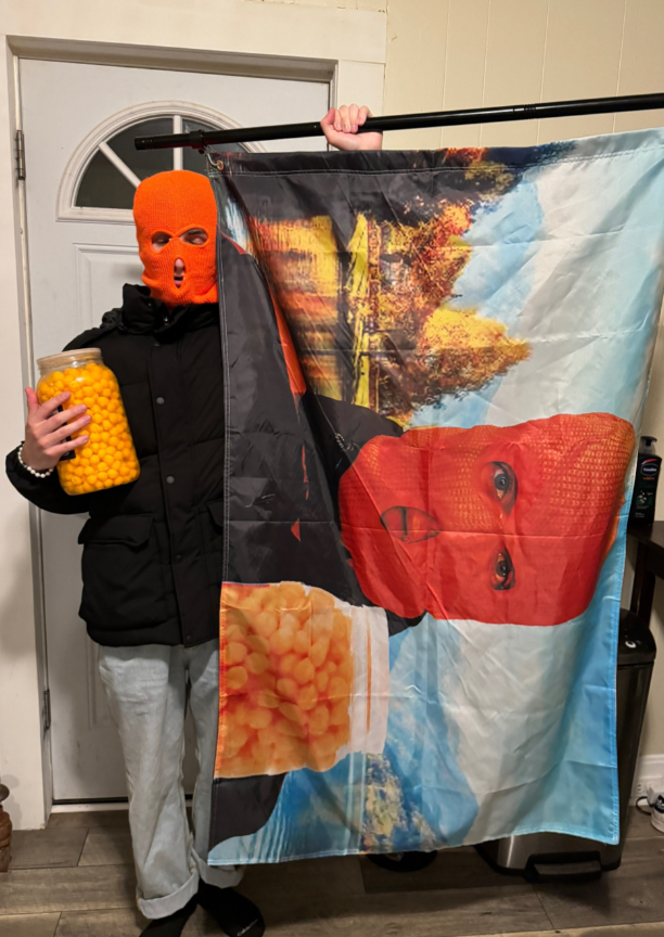 Person in a ski mask holds a jar and displays a large printed fabric with a stylized graphic face