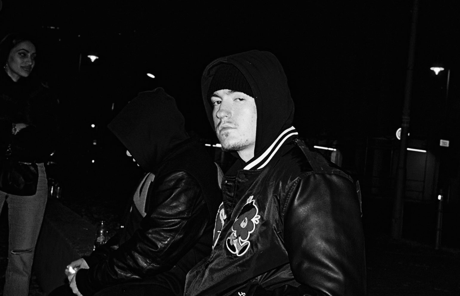 Individual in hood and jacket with embroidered details, leaning forward with a focused expression