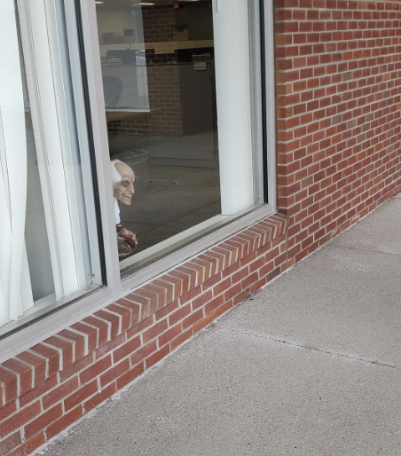 Elderly person doll peeking out with a smile from behind a partially open window on a brick building