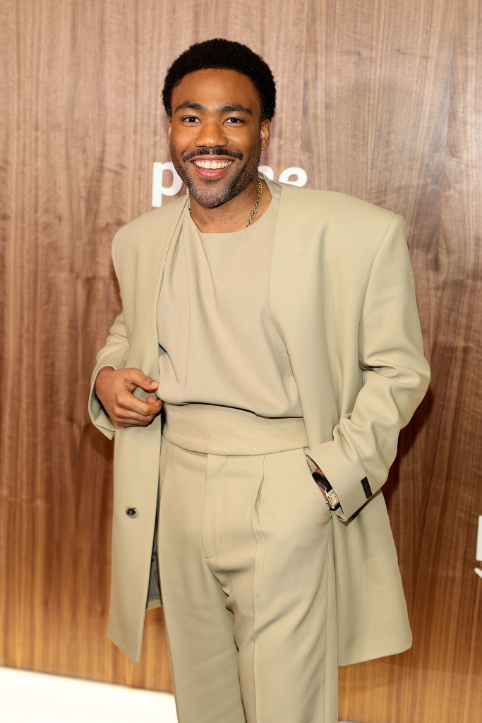 Donald Glover/Childish Gambino in a beige suit smiling at a premiere event with branded backdrop