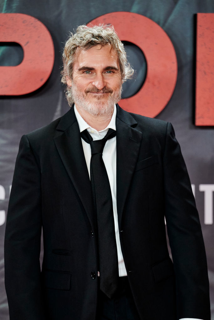Joaquin Phoenix in a black suit and white shirt, smiling at the camera, at an event