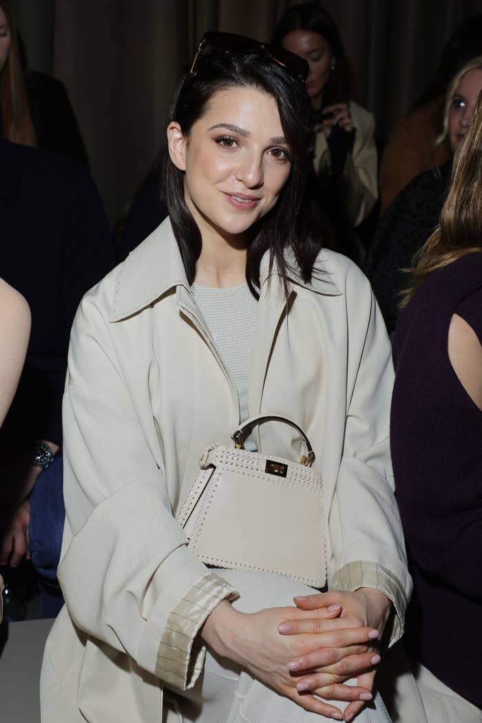 Marisa Abela sitting at an event wearing a light jacket over a textured top, accessorized with a small handbag