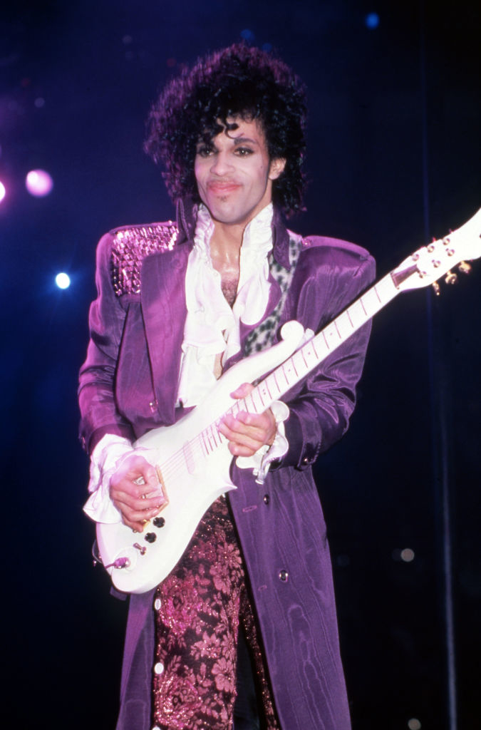 Prince performs onstage with a guitar, wearing a frilly shirt and purple jacket