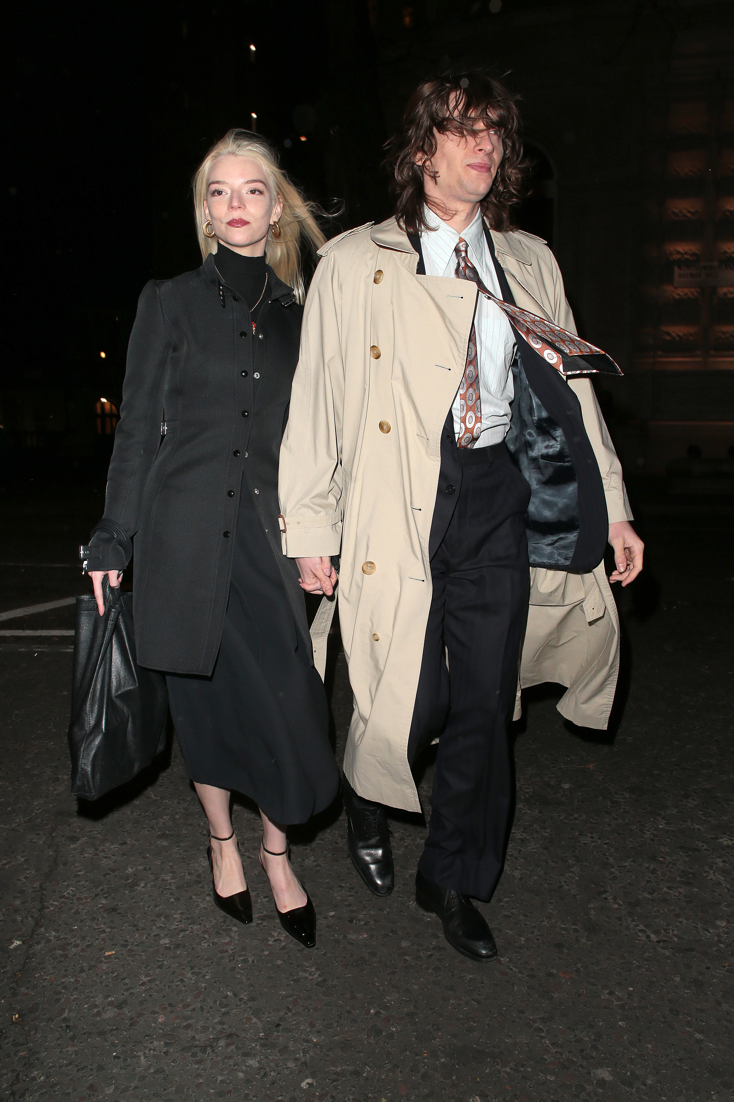 Two celebrities walking, the woman in a black dress and coat, the man in a beige trench coat and scarf, both holding bags
