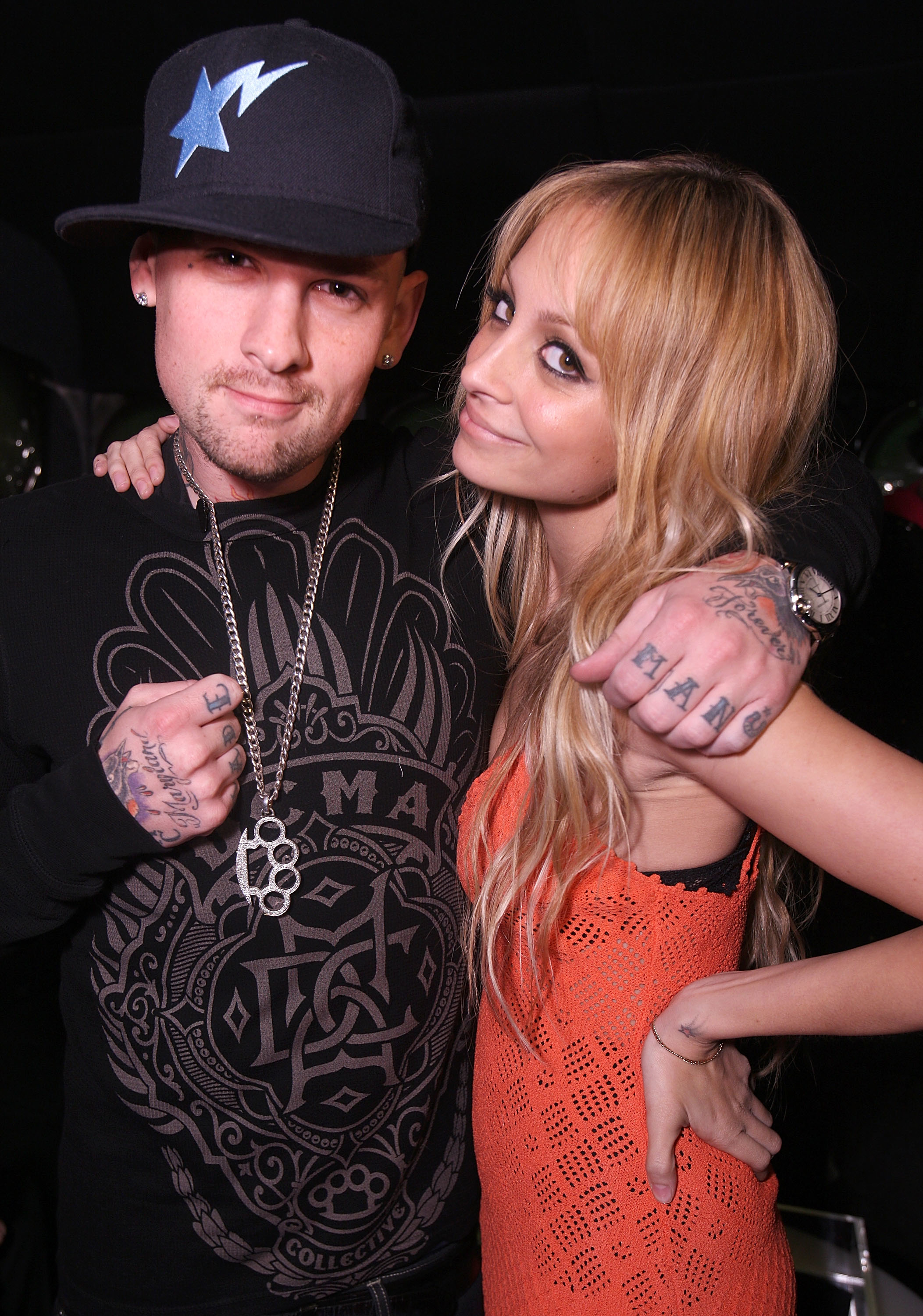 Two individuals posing closely; one wearing a graphic tee and cap, the other in a scoop neck top with visible tattoos