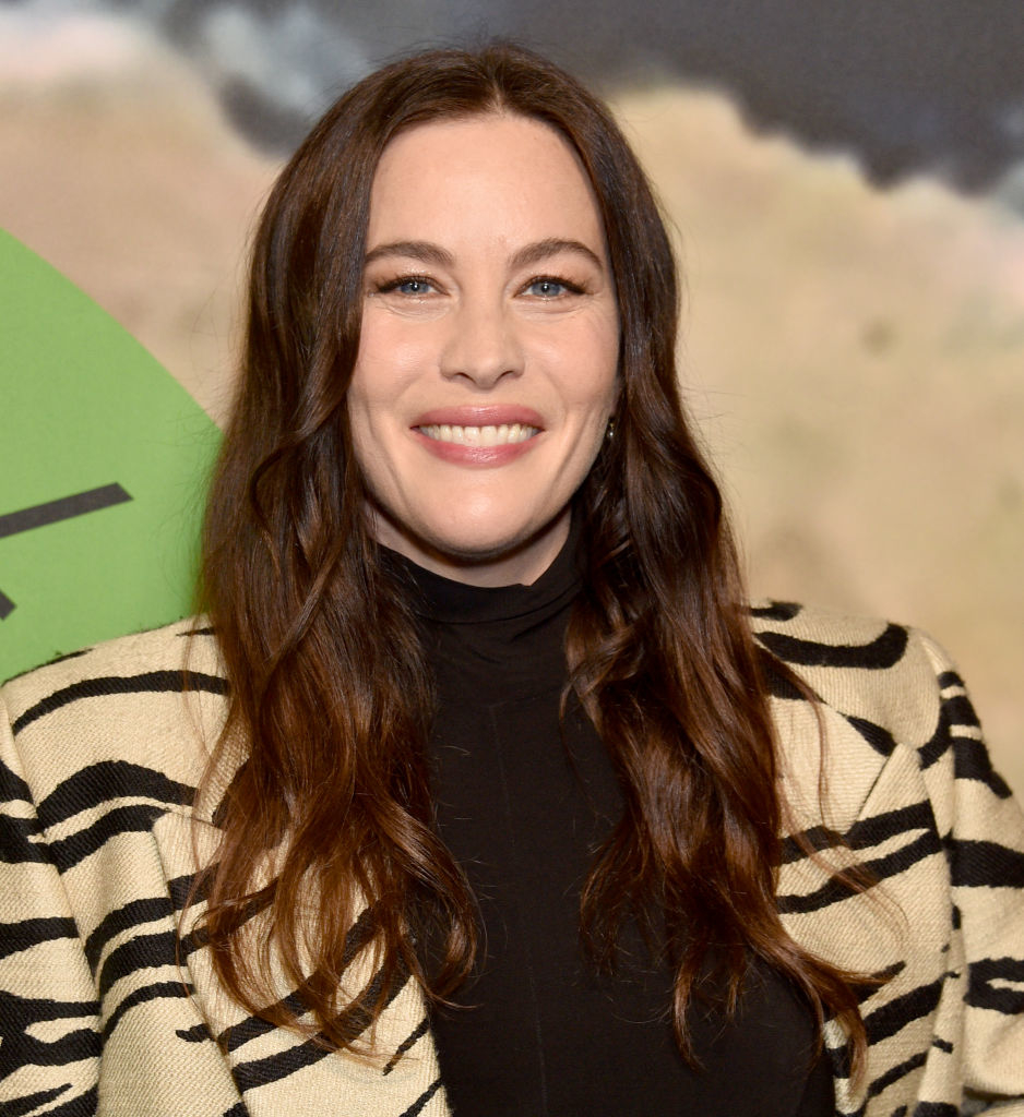 Liv Tyler with long hair wearing a striped blazer over a black top, smiling at a publicity event