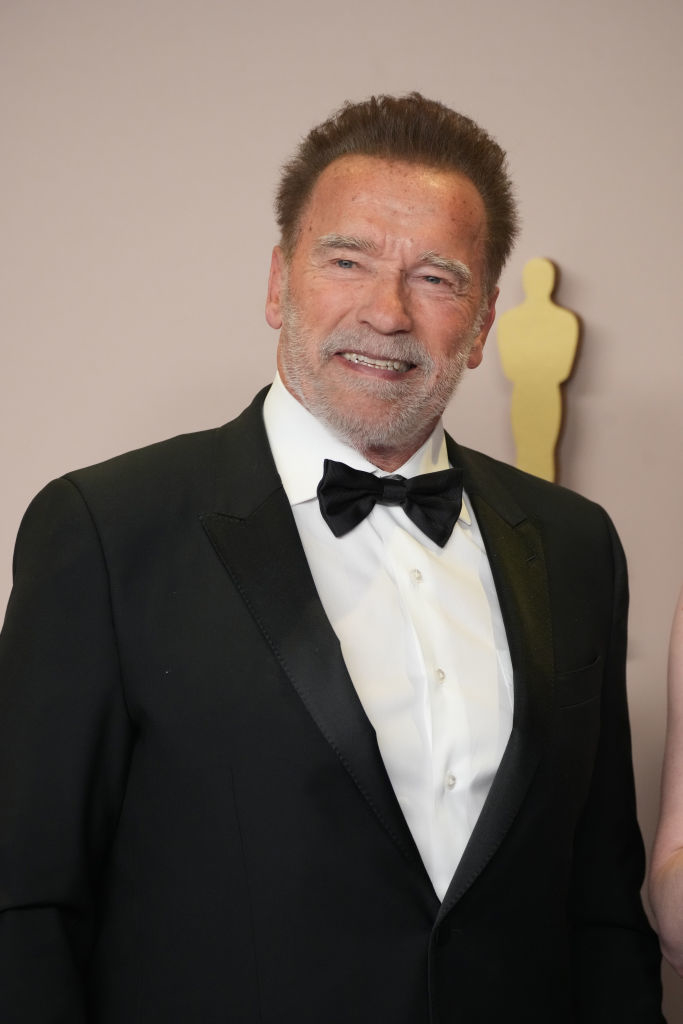 Arnold Schwarzenegger in a classic tuxedo with bowtie, standing before an award statuette