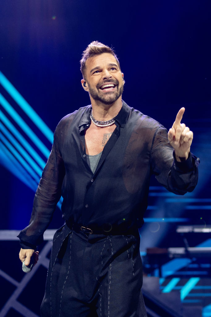 Ricky Martin performing on stage, wearing a semi-sheer black shirt and trousers, smiling and pointing upwards