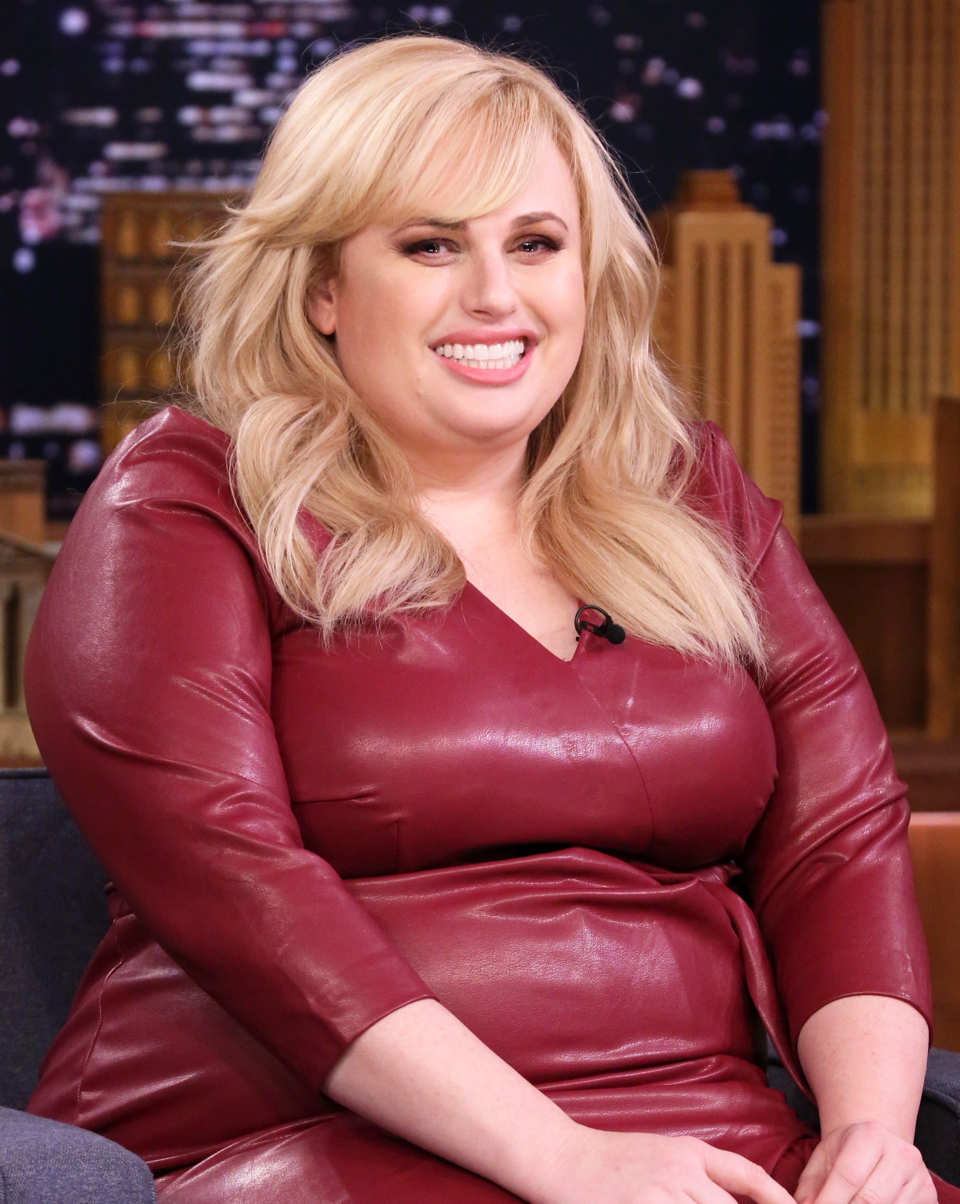 Rebel Wilson seated on a talk show set wearing a leather dress