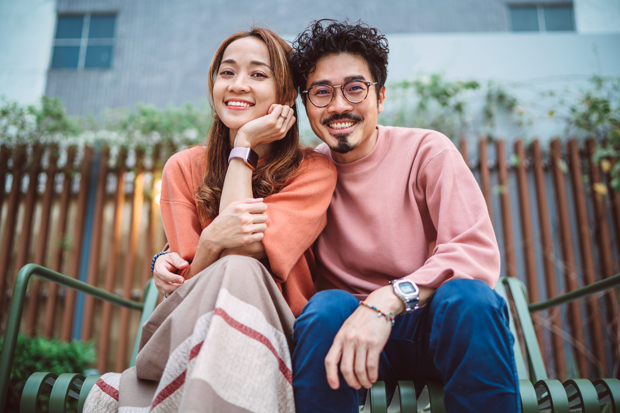Two people smiling and sitting closely together on a park bench, one resting their head on the other&#x27;s shoulder