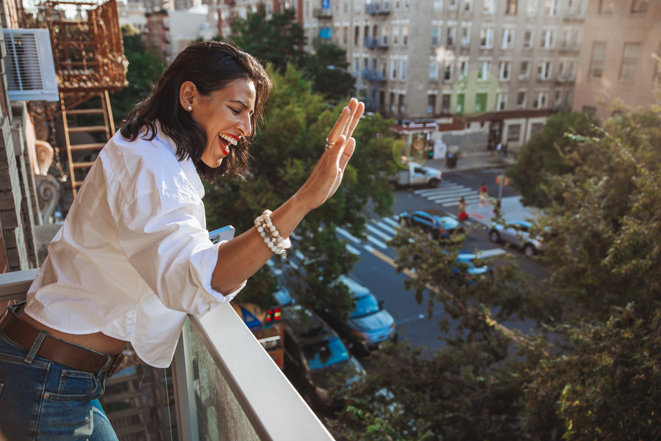 Woman on balcony waving to street below, wearing casual white blouse and jeans