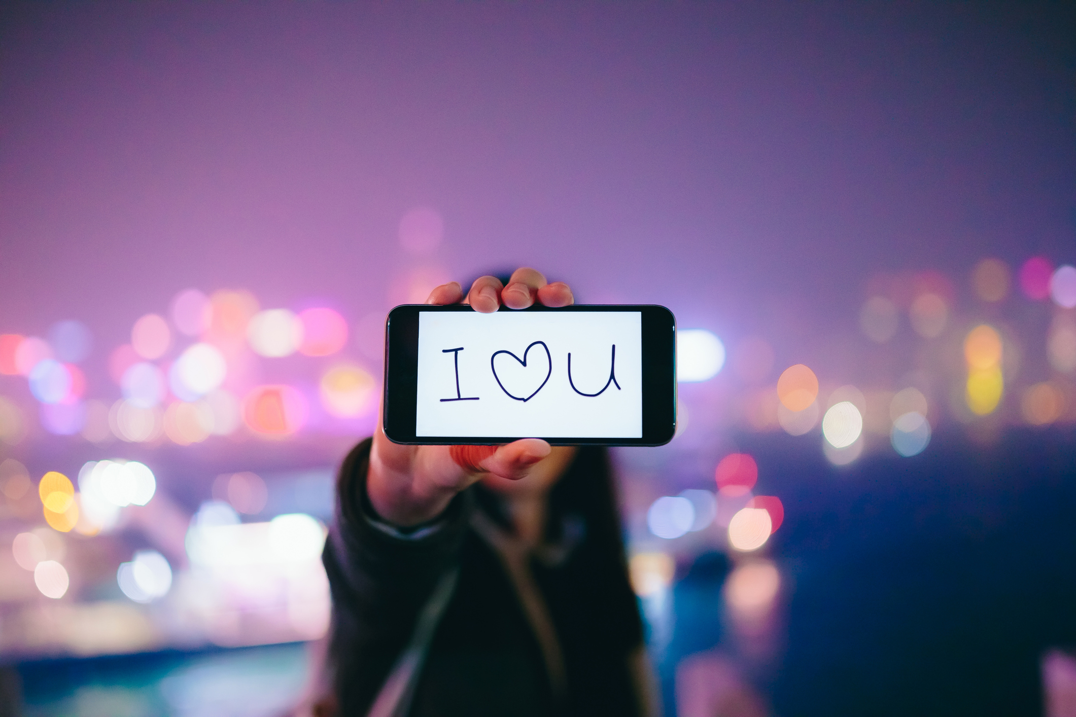 Hand holding smartphone with &quot;I ❤ U&quot; on screen, blurred city lights backdrop