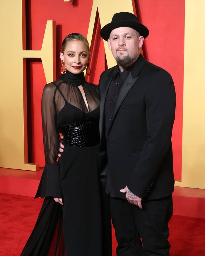 Two celebrities pose together; one in a sheer top and sequined dress, the other in a black suit and hat