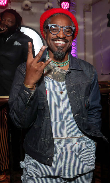 André 3000 in pinstripe suit and red hat making peace sign, smiling, at an indoor event