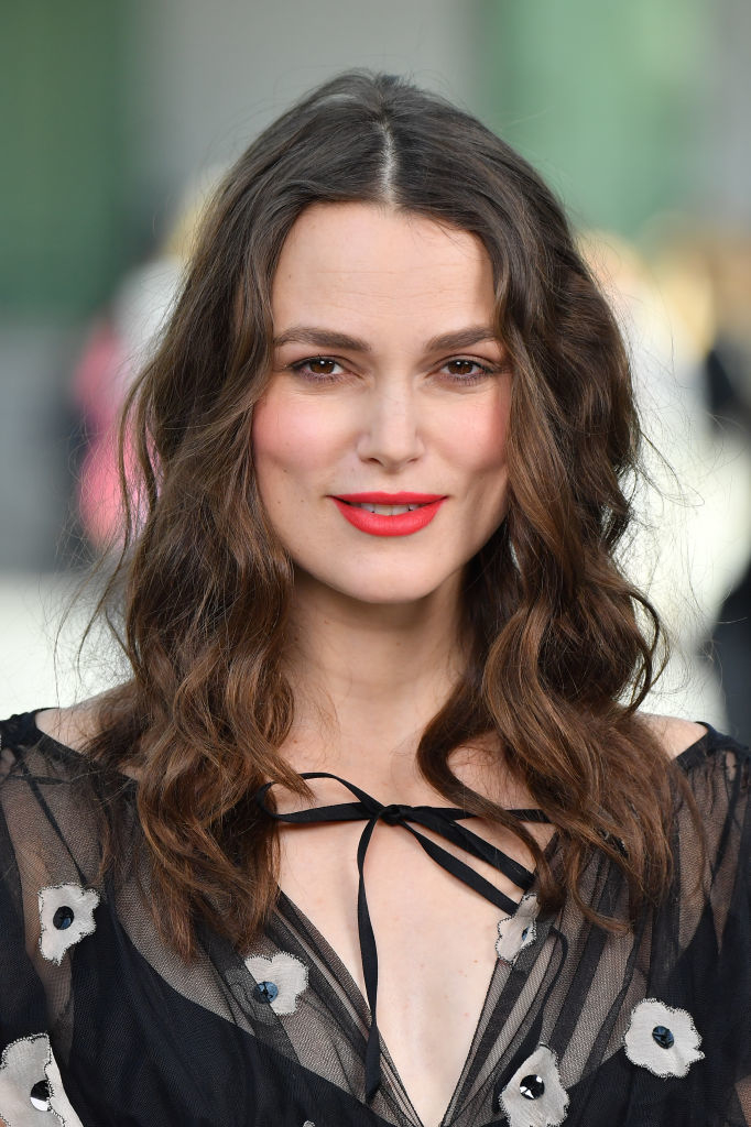 Keira Knightley in a sheer black outfit with floral accents, smiling at a camera