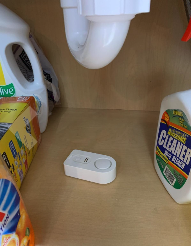 Laundry detergent and cleaning supplies on a shelf with a leak sensor placed below a drain pipe