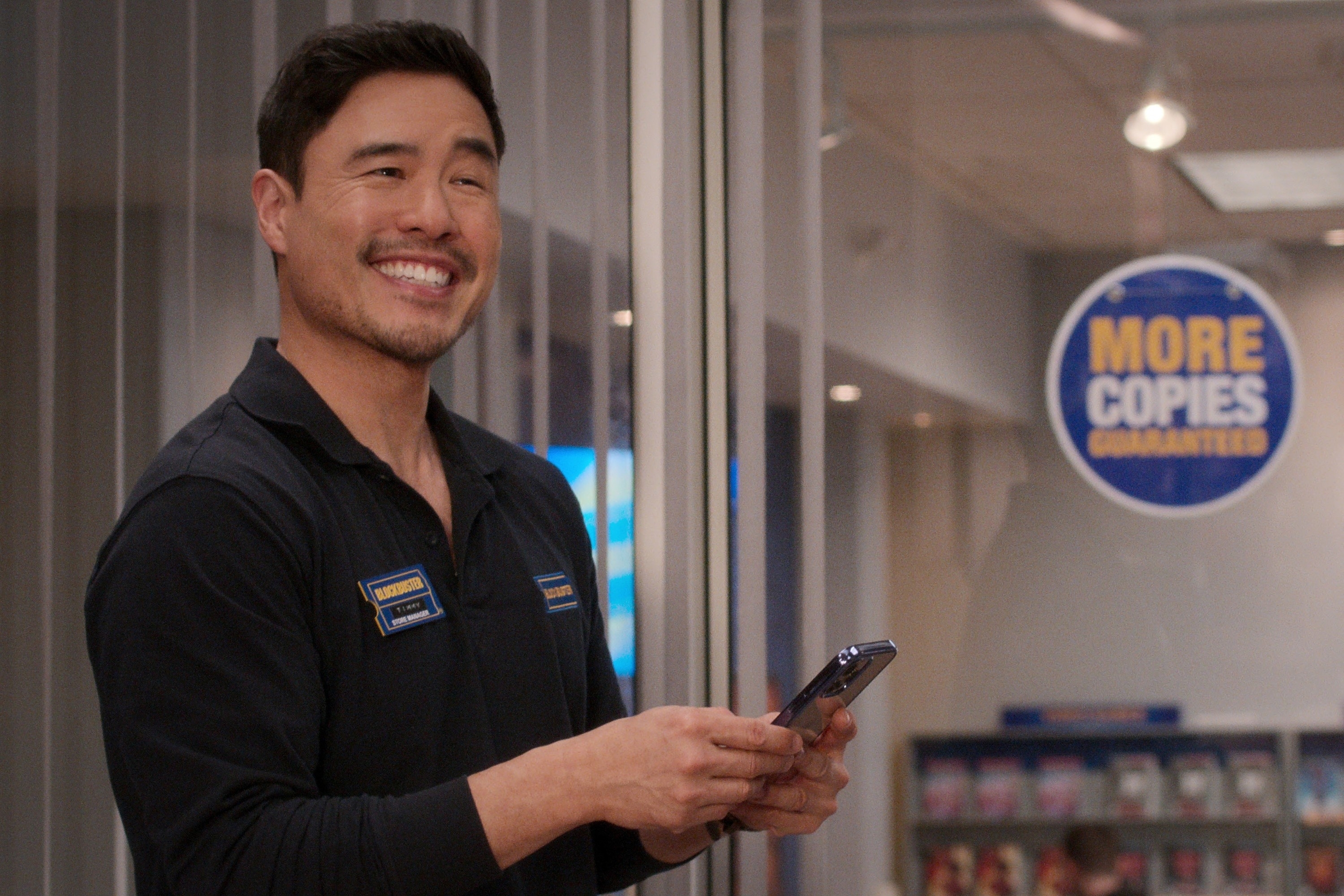 Randall Park in a scene in a store uniform smiling at his phone with a &quot;More Copies&quot; sign in the background