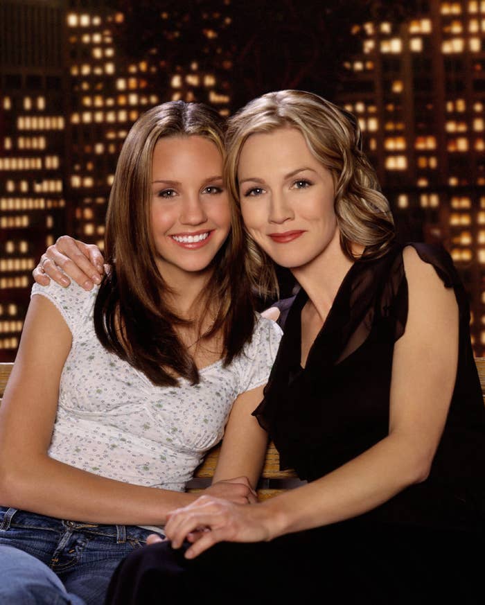 Jennie and Amanda Bynes embracing and smiling; one in casual top and jeans, the other in a black dress. They pose before a cityscape backdrop