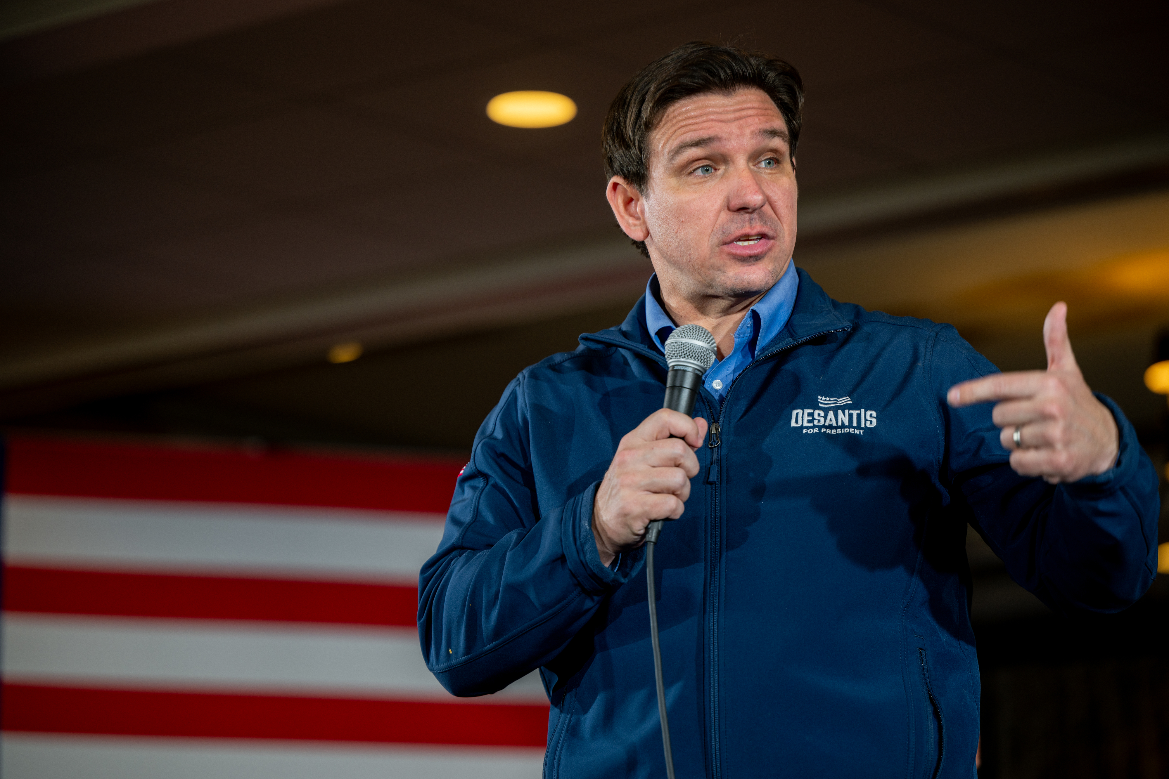 Ron DeSantis in a branded jacket holding a microphone, speaking in front of an American flag