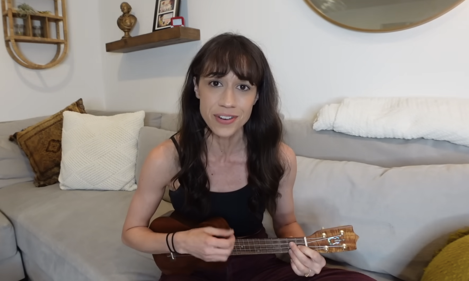 Woman smiling, holding a ukulele, sitting on a couch with cushions and a trophy in the background