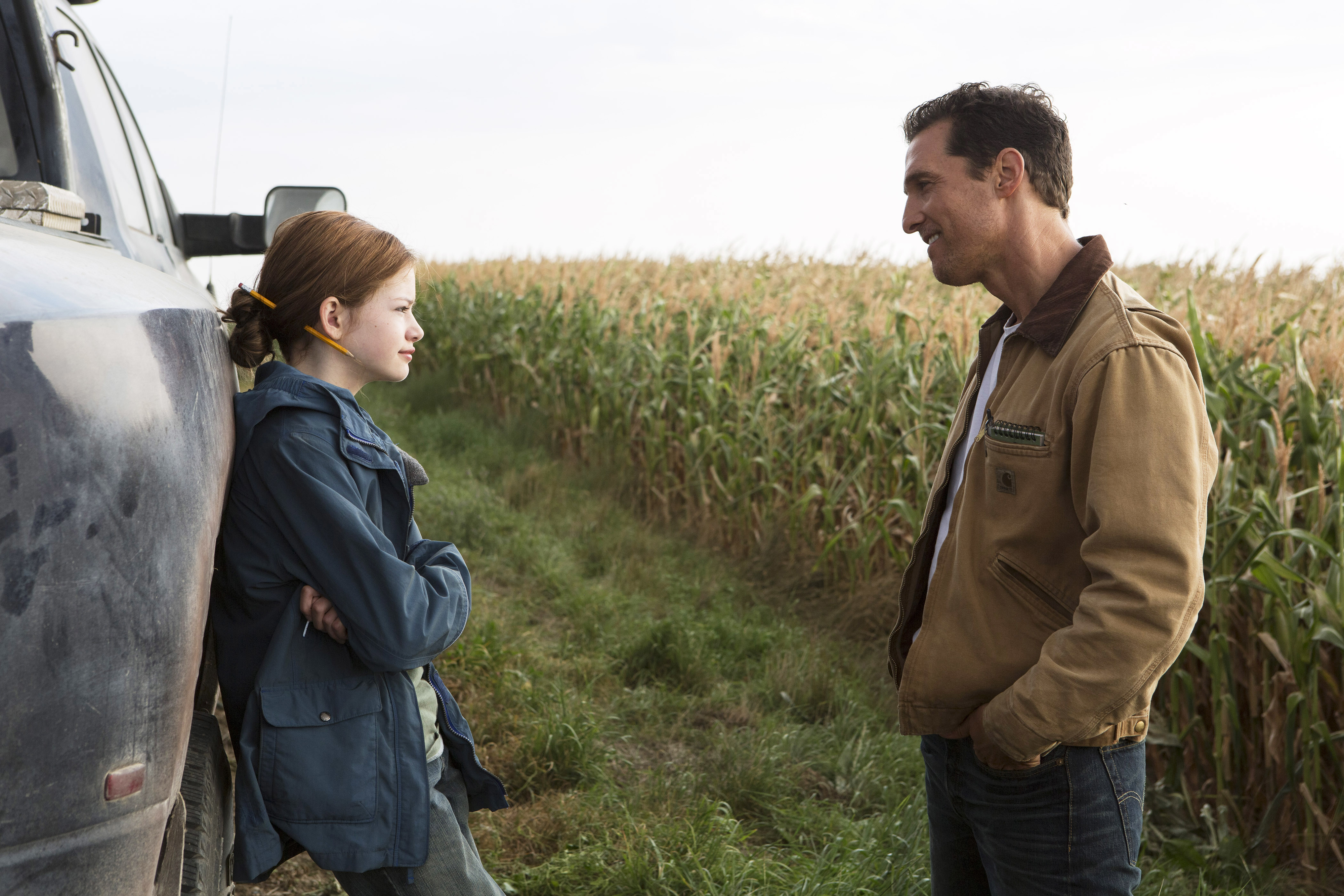 A man and a young girl stand talking next to a truck in a cornfield