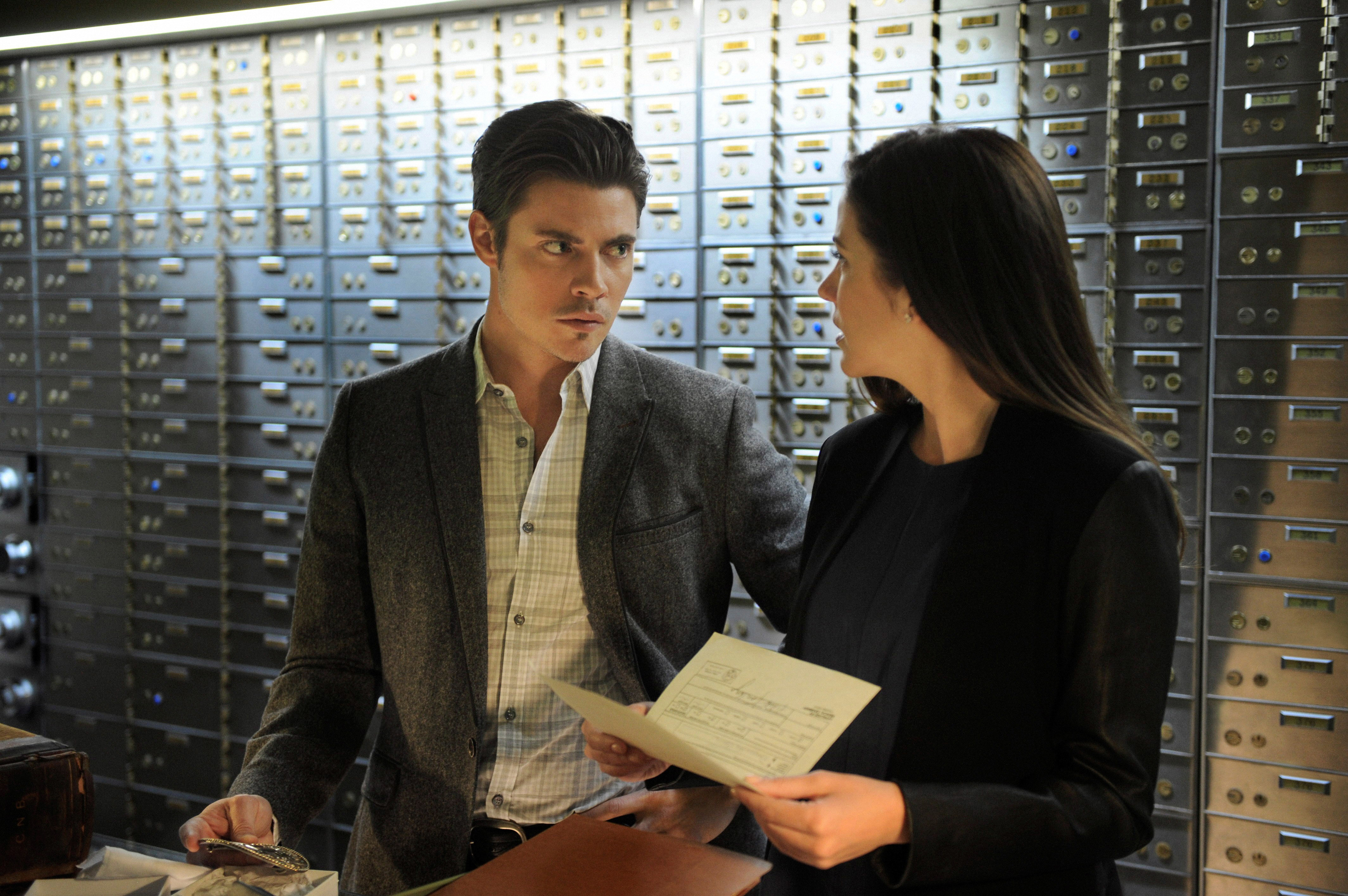Josh Henderson and Julie Gonzaloin a scene, standing in front of a wall of safety deposit boxes, engaged in discussion