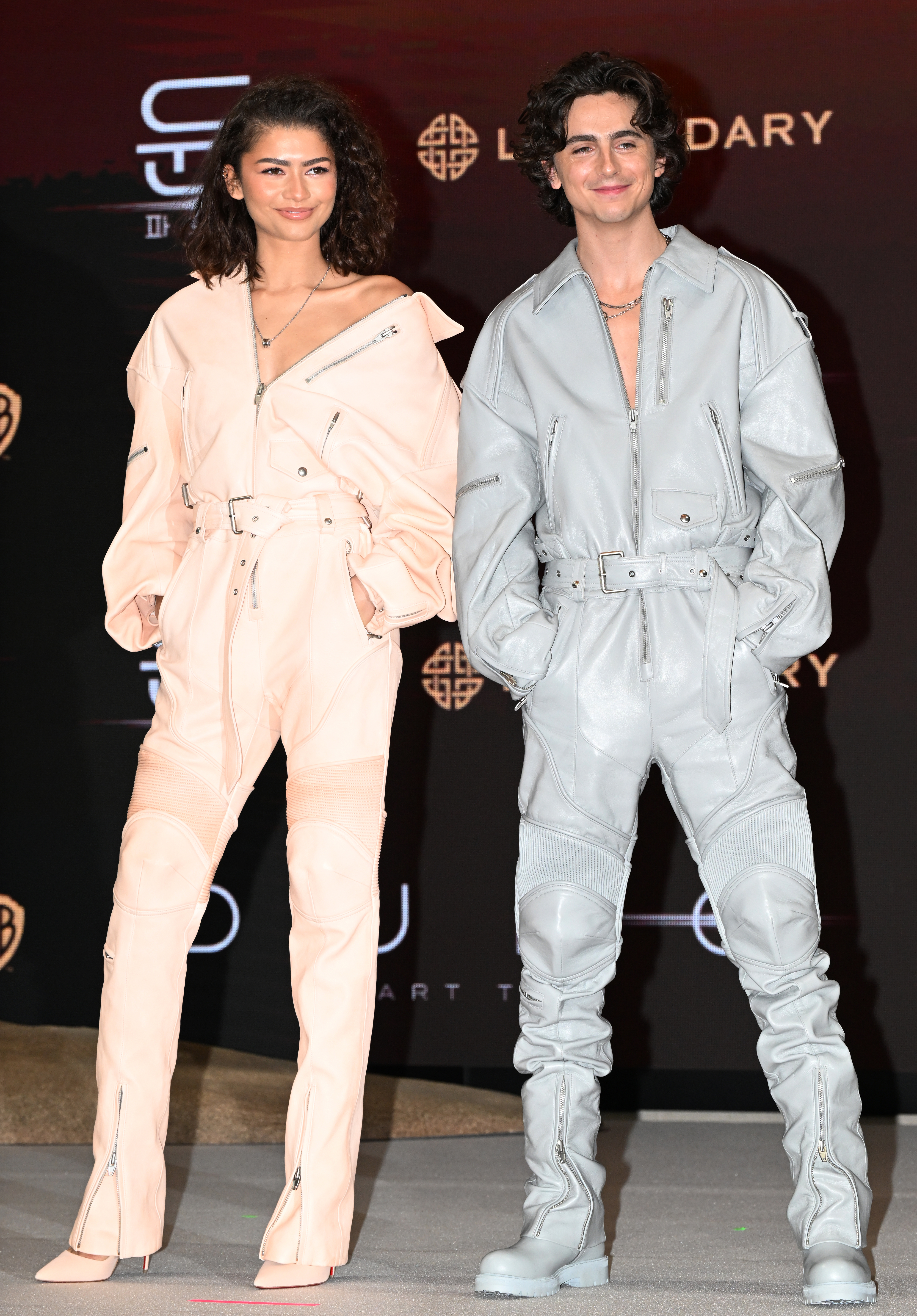 The costars posing in matching belted jumpsuits and tall boots at an event