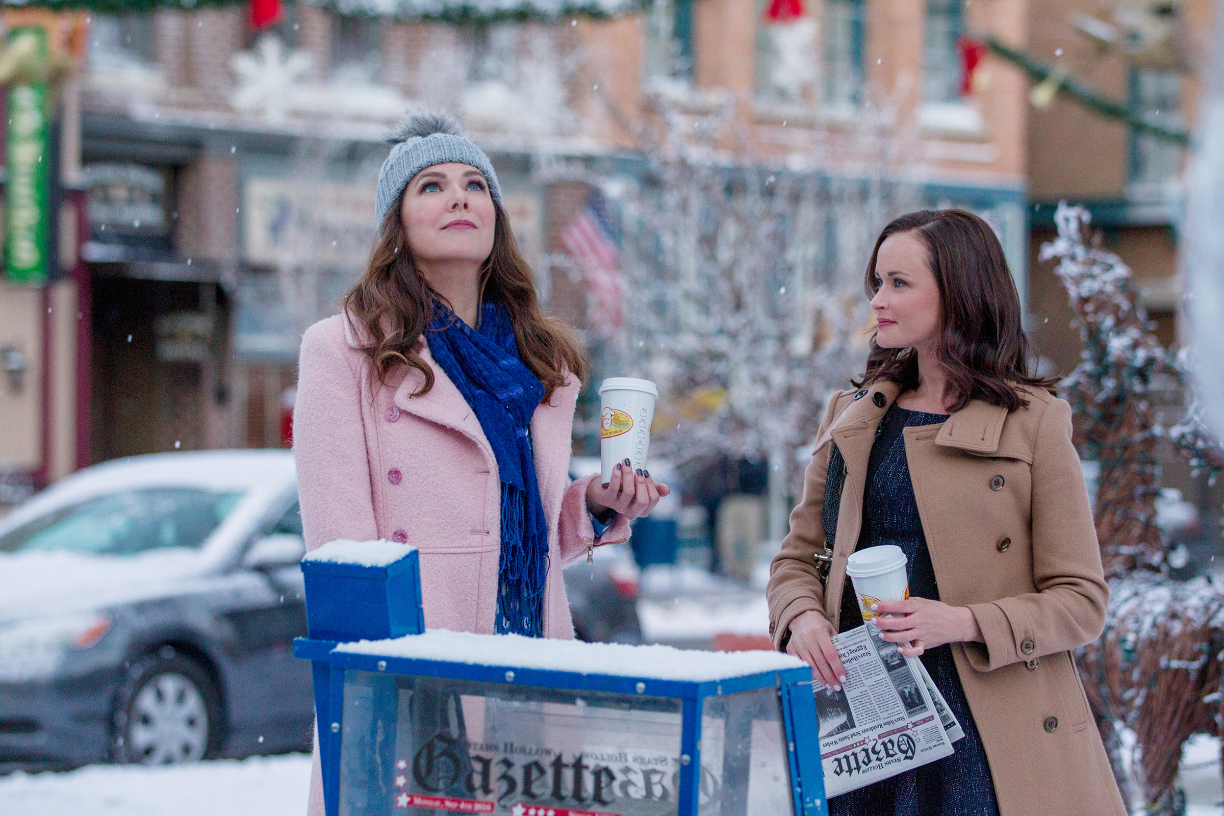 Lorelai and Rory stand outside with coffee cups, winter attire, and a snowy backdrop