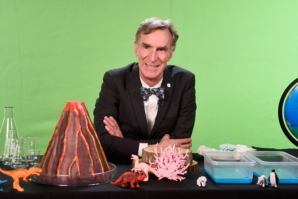 Bill Nye with a bowtie, leaning on a table with educational science display items