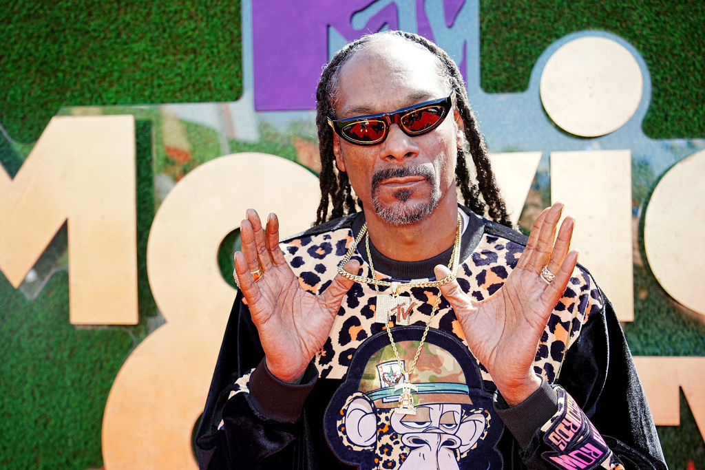 Snoop Dogg posing with hands together at an event, wearing a patterned jacket, sunglasses, and a large pendant
