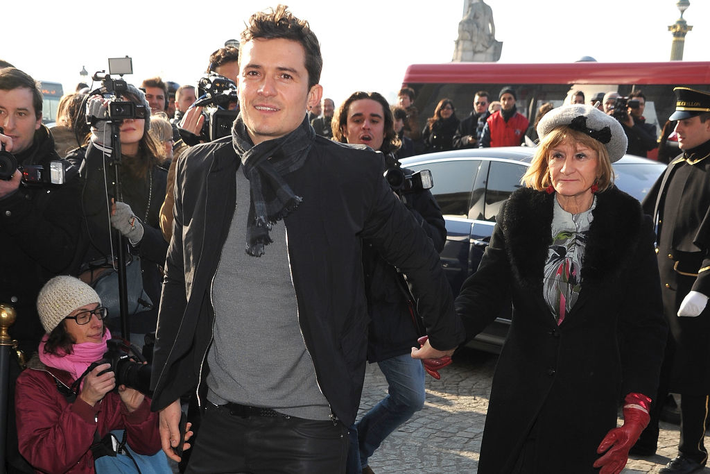 Orlando Bloom in a casual jacket and scarf holding hands with an elderly woman in a coat and hat, surrounded by photographers