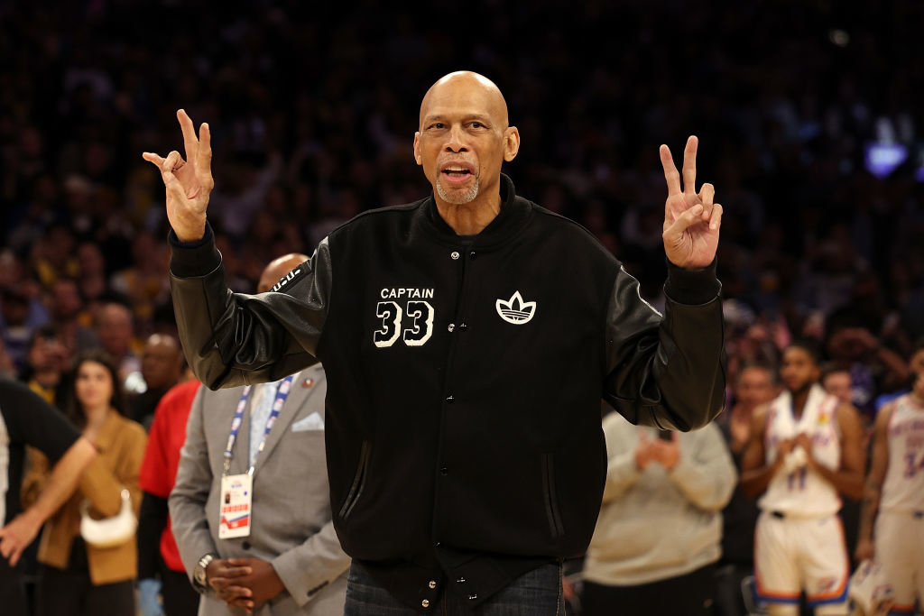 Kareem Abdul-Jabbar on a basketball court, wearing a black &#x27;CAPTAIN 33&#x27; jacket, gesturing peace signs with both hands