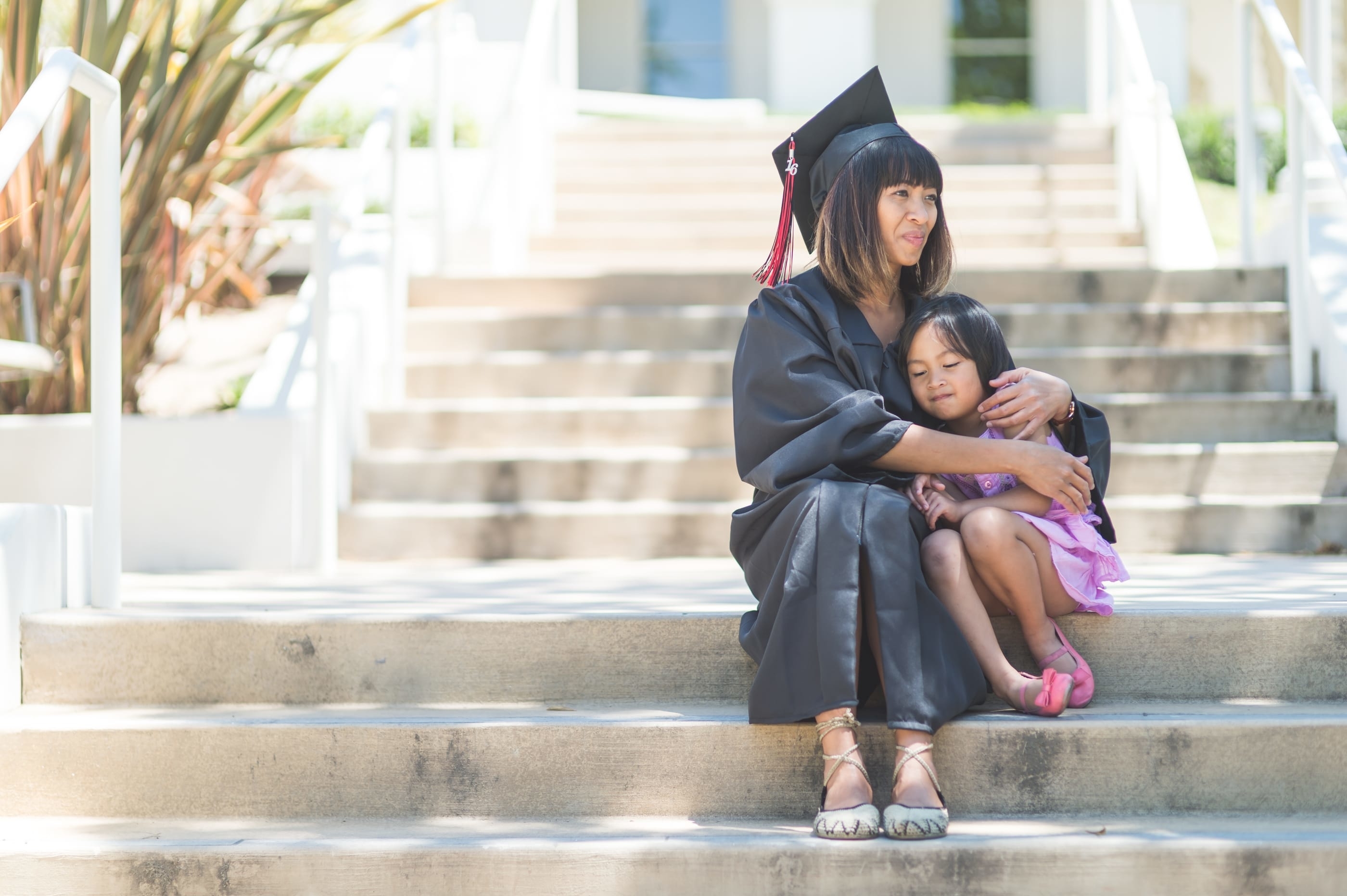 Graduate in cap and gown sitting on steps, embracing a young child