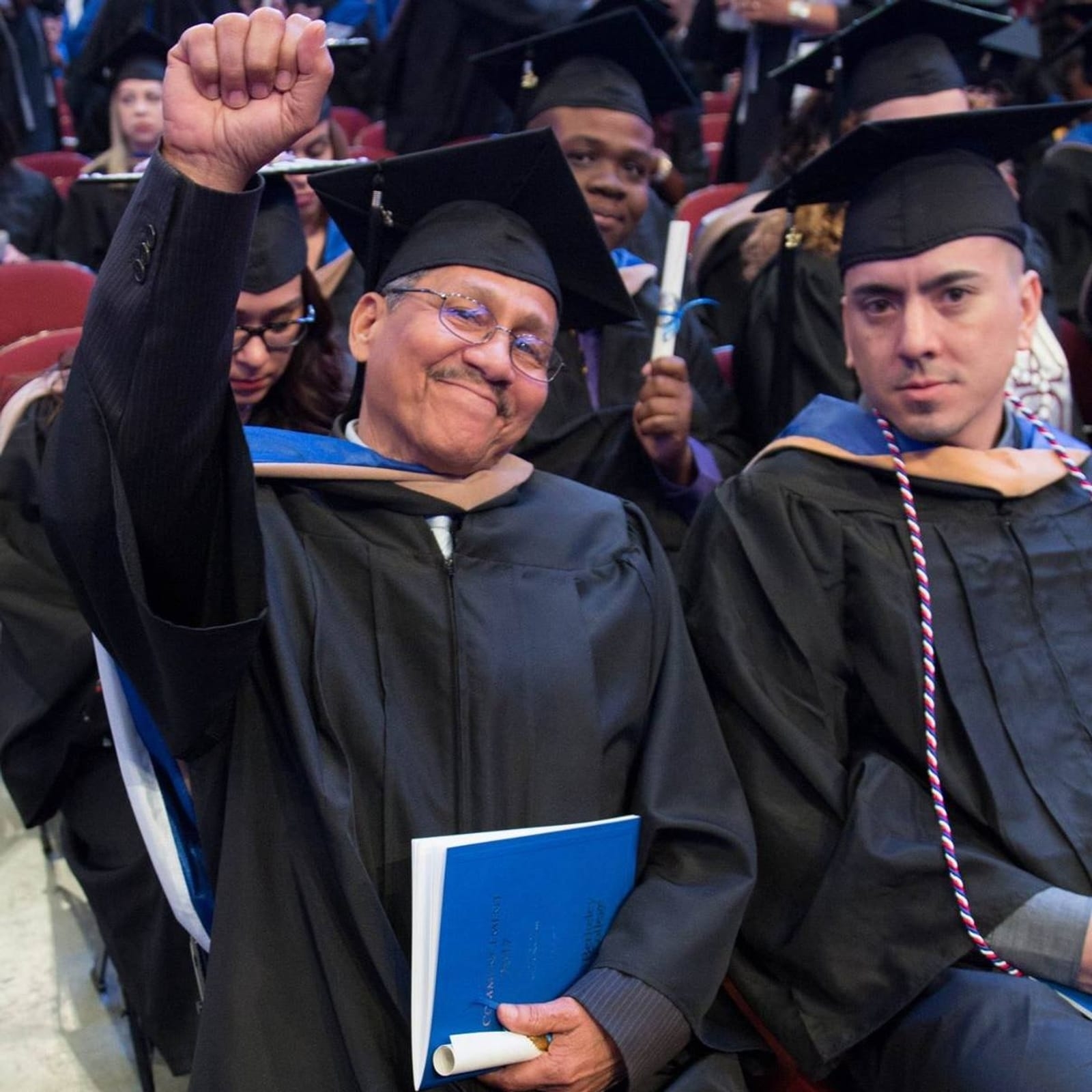 Graduate in cap and gown raises fist triumphantly at graduation ceremony, surrounded by fellow graduates