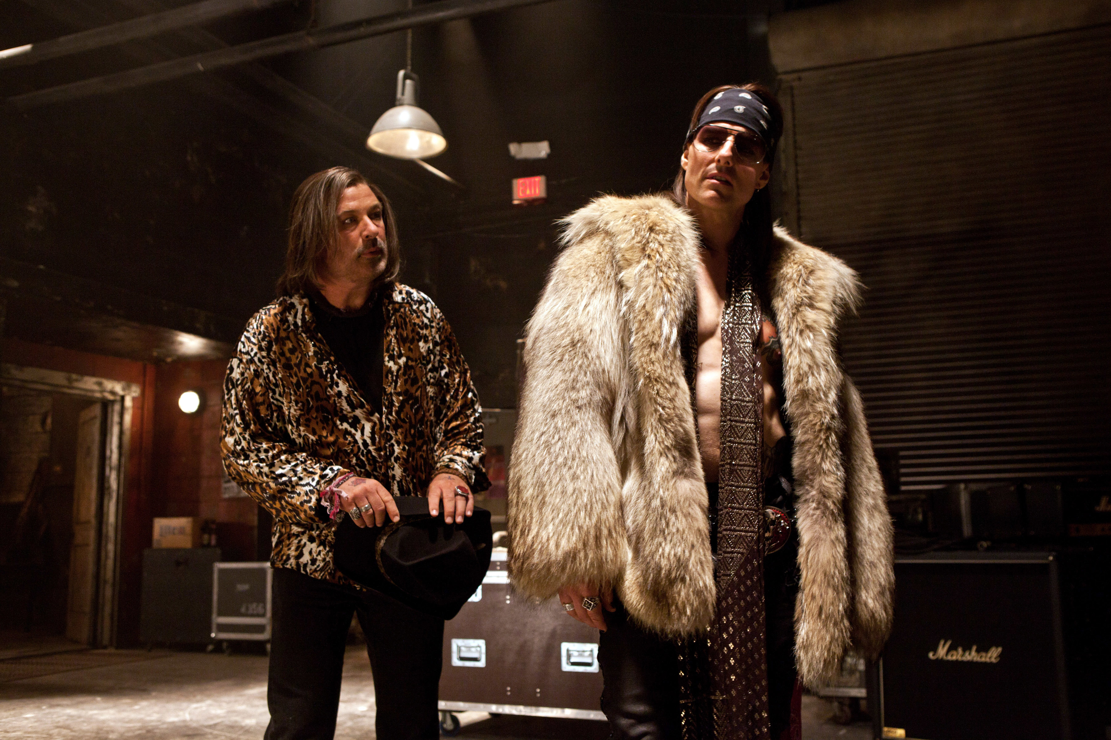 Alec Baldwin and Tom Cruise on a set dressed as rock stars with leather and fur clothing, standing near musical equipment
