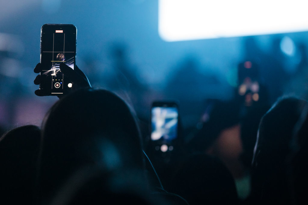 Audience with phones raised at a concert, capturing the event. Focus on one phone&#x27;s screen showing the stage