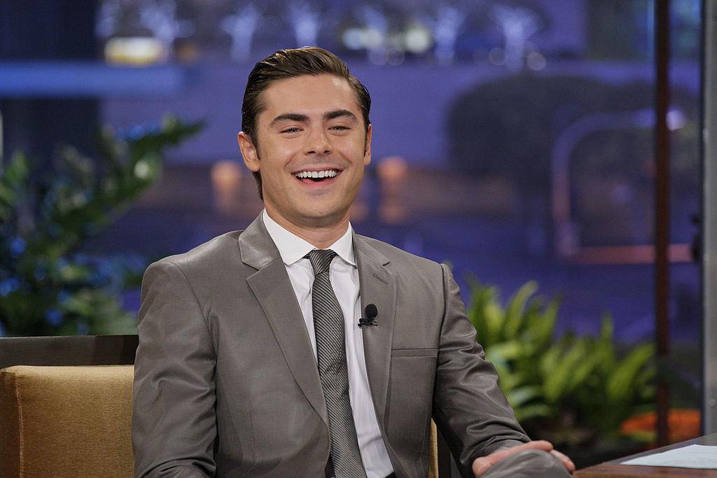 Zac Efron wearing a gray suit and tie, smiling during a TV interview