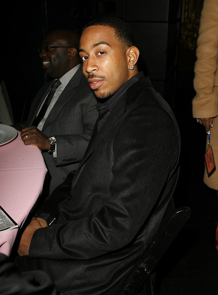 Ludacris seated at an event, wearing a black shirt and blazer, with a serious expression