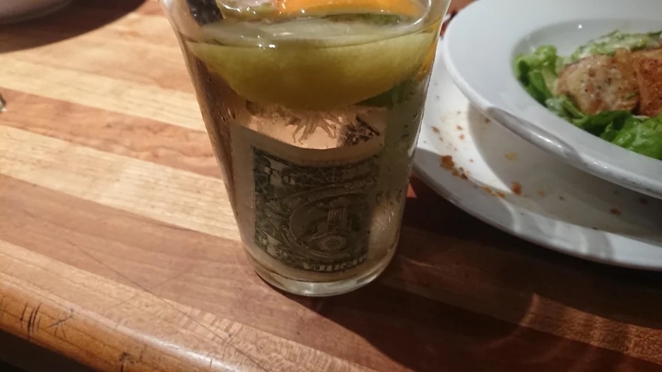 A glass of water with a one dollar bill submerged inside, next to a salad plate on a wooden table
