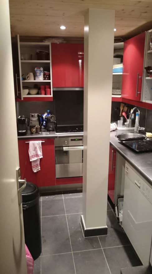 A kitchen with appliances and cabinets, pillar in center, trash can on left, dish towel on right