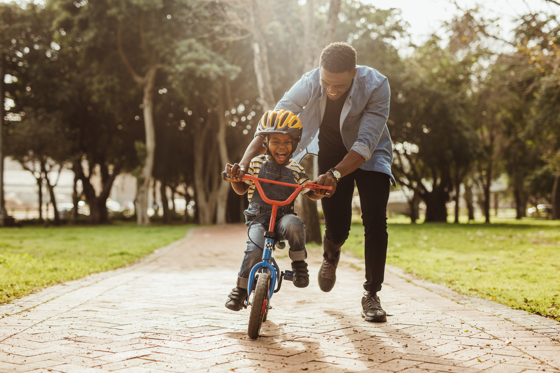 Adult assisting a child with learning to ride a bike in a park setting. Both are smiling