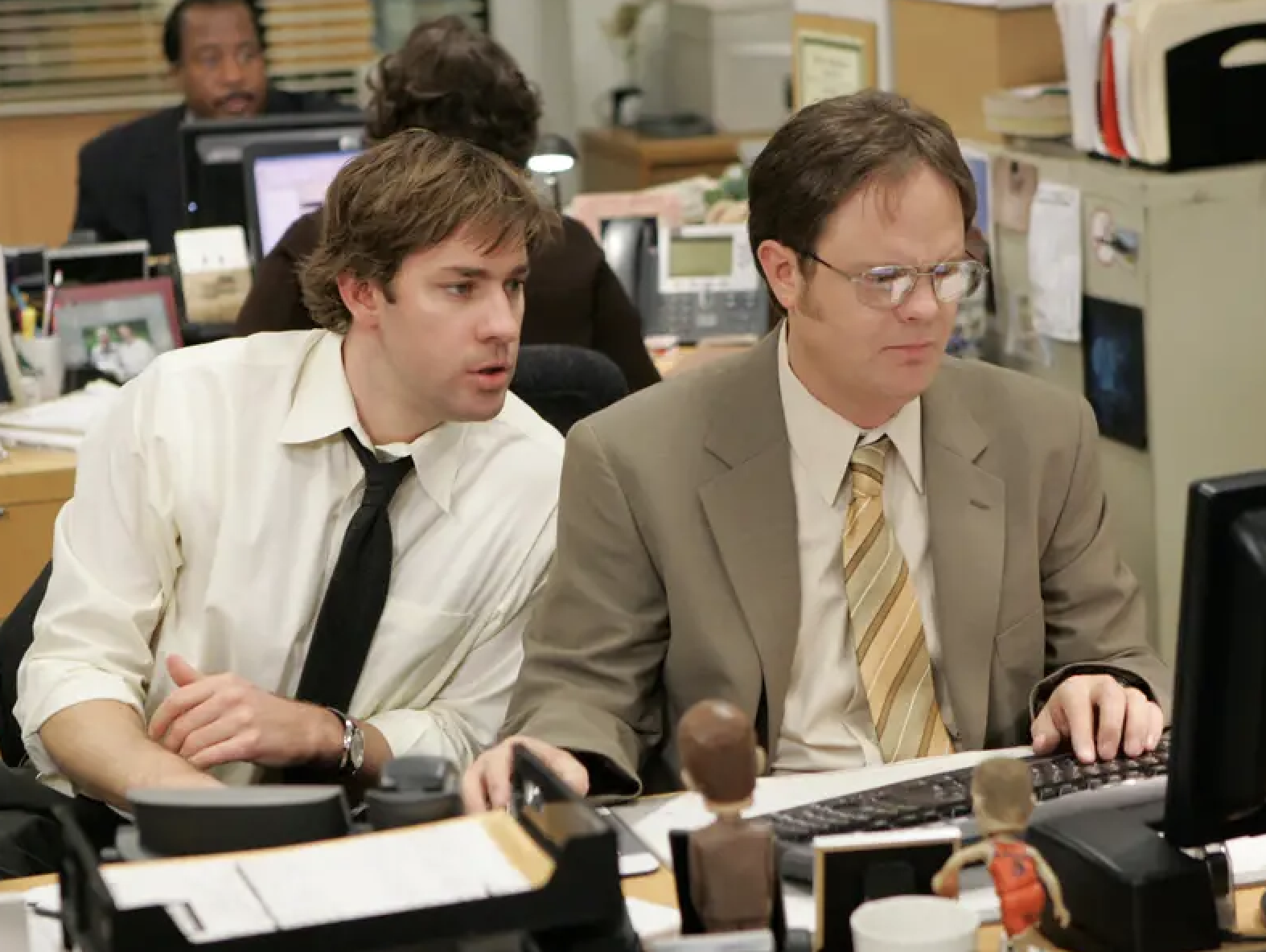 Two male characters from The Office, Jim and Dwight, seated at desks, appear to be working and discussing