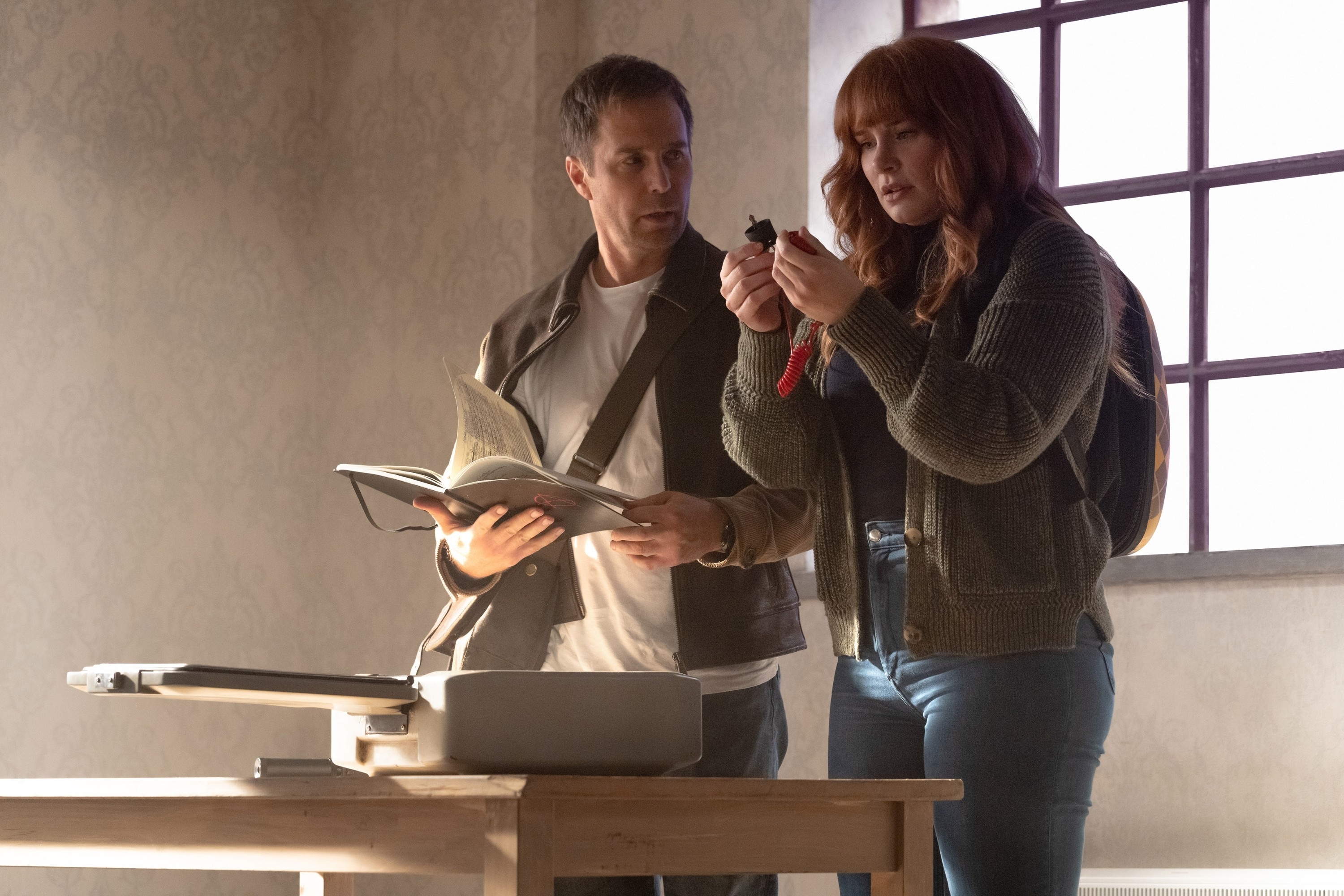 in a scene, Sam Rockwell holding book and pen with a focused expression, Bryce Dallas Howard examining a small object, dressed in casual attire