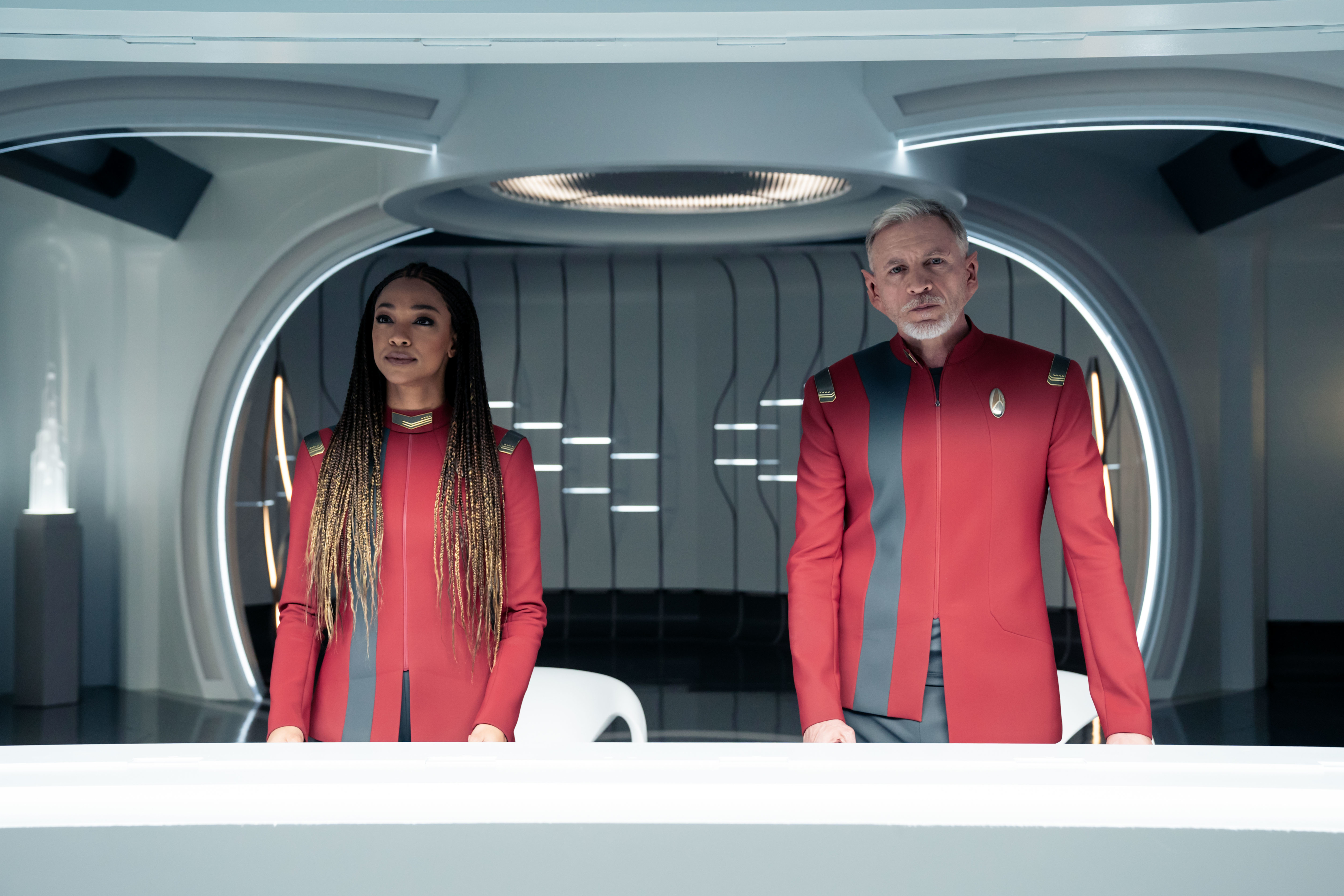Two actors in a sci-fi show, dressed in futuristic red uniforms, standing on a spaceship bridge