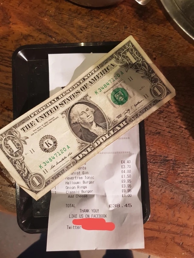 A person&#x27;s hand placing a US dollar bill on a restaurant receipt with a total amount written in British pounds